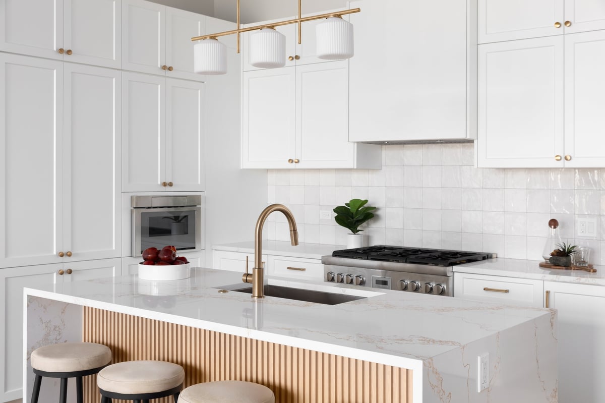A kitchen detail with white cabinets and square tile backsplash, a waterfall marble countertop and wood slat panel front, and a gold light fixture, hardware, and faucet.