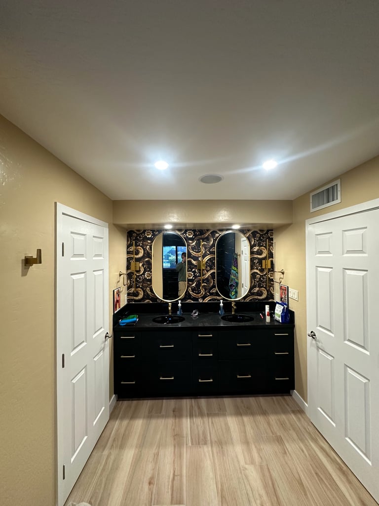 Modern bathroom with black vanity, dual oval mirrors, decorative black and gold tile backsplash, LED ceiling lights, and white doors on beige walls.