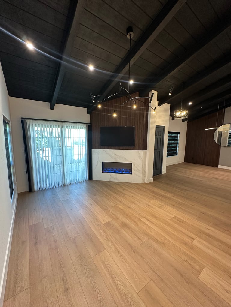 Modern open-concept living space with wooden flooring, white fireplace accent wall, dark ceiling beams, pendant lighting, and glass doors with vertical blinds