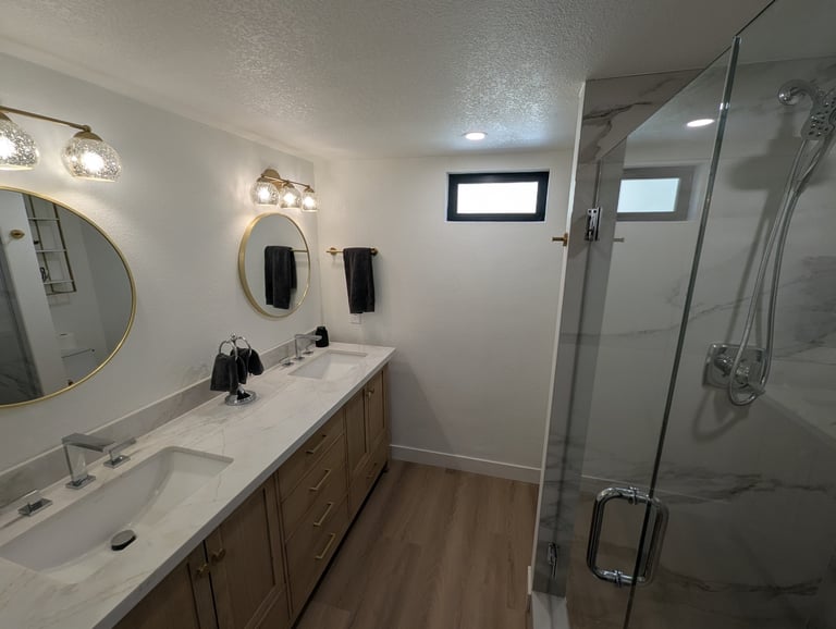 Modern bathroom with double vanity sink, round mirrors, wall sconces, wood-look flooring, and frameless glass shower enclosure