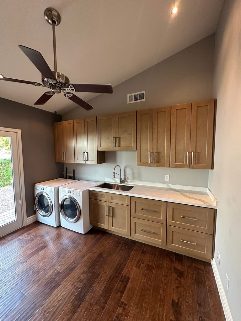 Modern laundry room with wood cabinets, two front-load washers, sink counter, and ceiling fan overhead.