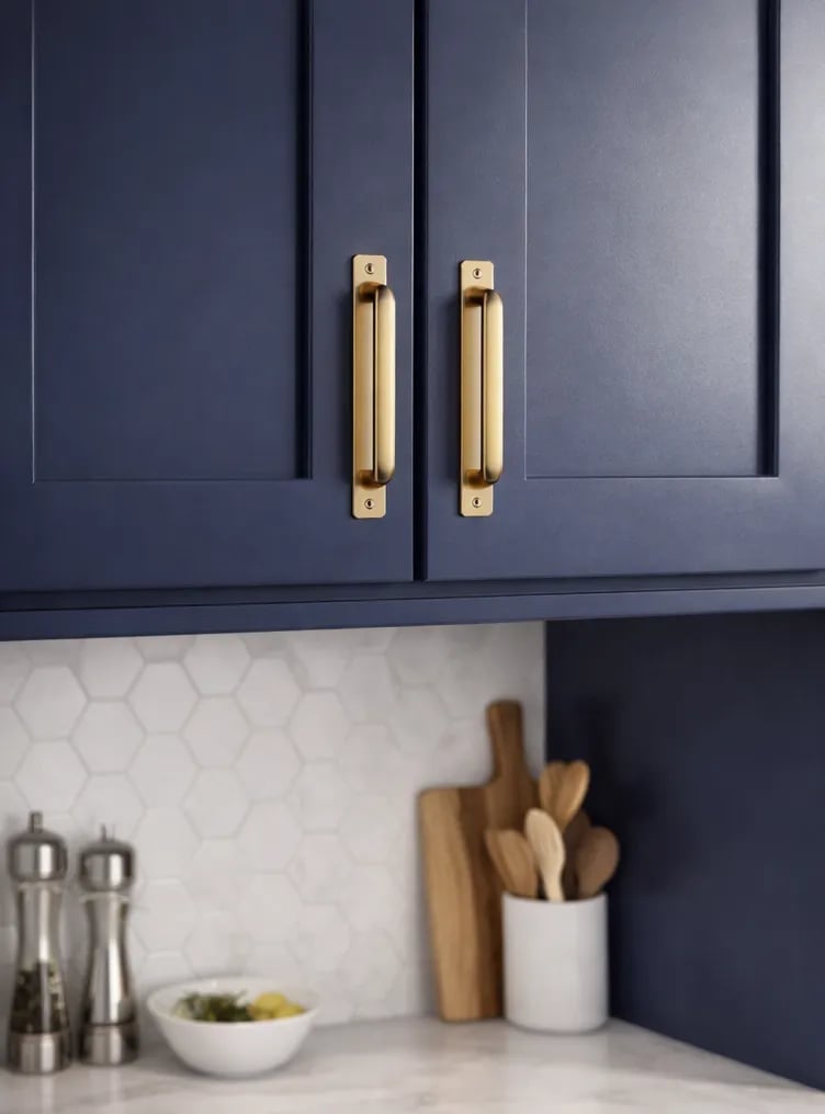 Dark blue kitchen cabinet with brass handles above white hexagonal tile backsplash and countertop with cooking utensils